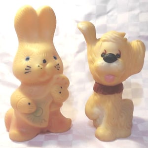 May include: Two vintage rubber toys: a light orange bunny holding a rattle and a light orange dog with a red collar. The bunny has blue eyes and black whiskers. The dog has a black nose and a pink tongue.