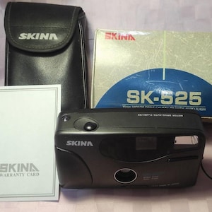 May include: A black and silver SKINA SK-525 35mm film camera with a built-in flash. The camera is in a black leather case and comes with a warranty card.