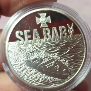 Peut inclure: Une pièce de monnaie circulaire de couleur argentée enfermée dans une capsule de protection en plastique transparent. La pièce présente un relief de bateau sur l'eau avec le texte SEA BABY au-dessus et un emblème en forme de croix au centre.