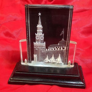 May include: A clear glass rectangular display featuring a detailed etched image of a building with a tall tower and a star on top. The display is set on a black base with clear supports.