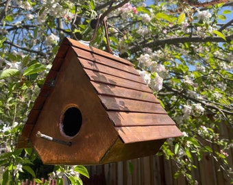 Puede incluir: Una casa para pájaros de cobre con un techo triangular de paneles metálicos horizontales, un orificio de entrada circular y una pequeña percha de madera que sobresale. La estructura cuelga de un gancho metálico frente a un árbol en flor.