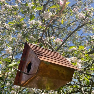 Puede incluir: Una casa para p&aacute;jaros de cobre con un techo inclinado y acanalado, que presenta un orificio de entrada circular con una peque&ntilde;a percha de madera que sobresale. La estructura est&aacute; suspendida por un alambre met&aacute;lico y tiene un acabado brillante.