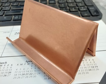 Puede incluir: Un soporte de exhibición de metal color cobre con un perfil triangular y una repisa frontal. El soporte presenta un acabado metálico liso y está diseñado para sostener objetos en posición vertical. Tiene una construcción angular simple.