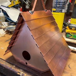 Puede incluir: Una casa para p&aacute;jaros triangular construida con l&aacute;minas de cobre pulido. El techo presenta paneles horizontales superpuestos y el panel frontal incluye un orificio de entrada circular y un orificio de drenaje m&aacute;s peque&ntilde;o. Un asa de metal est&aacute; fijada en la parte superior.