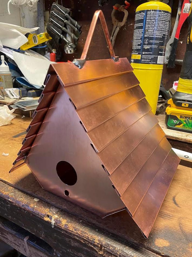 Puede incluir: Una casa para p&aacute;jaros triangular construida con l&aacute;minas de cobre pulido. El techo presenta paneles horizontales superpuestos y el panel frontal incluye un orificio de entrada circular y un orificio de drenaje m&aacute;s peque&ntilde;o. Un asa de metal est&aacute; fijada en la parte superior.
