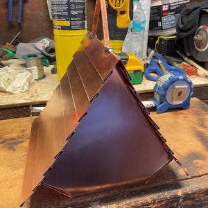 Puede incluir: Una estructura triangular de cobre con paneles met&aacute;licos superpuestos y un asa central en la parte superior. El objeto tiene un acabado met&aacute;lico pulido con costuras visibles donde se conectan los paneles, creando una forma geom&eacute;trica puntiaguda.