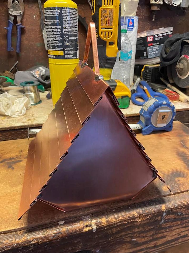 Puede incluir: Una estructura triangular de cobre con paneles met&aacute;licos superpuestos y un asa central en la parte superior. El objeto tiene un acabado met&aacute;lico pulido con costuras visibles donde se conectan los paneles, creando una forma geom&eacute;trica puntiaguda.
