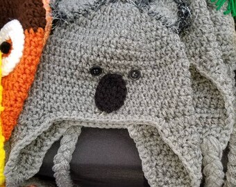 May include: A gray crocheted koala hat with button eyes and a black nose. The hat features ear flaps and a fuzzy trim around the face. Other colorful crocheted animal hats are visible in the background.
