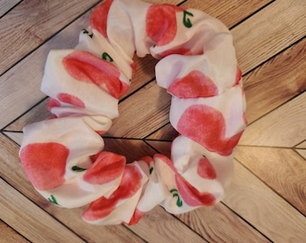 May include: A white scrunchie with a repeating pattern of red and pink fruit shapes and small green leaves. The fabric is gathered, creating a textured, circular accessory. This hair tie is suitable for various hairstyles.