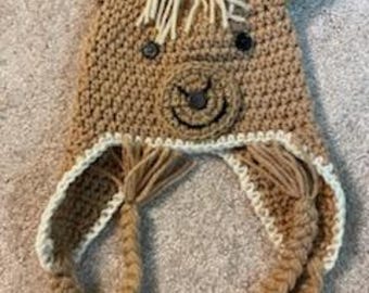 May include: A handmade crochet horse hat in shades of brown and cream. The hat features a horse face with a smiling mouth, button eyes, and a fringe mane. Braided tassels hang from the sides.