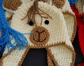 May include: Cream-colored crocheted hat with brown accents, featuring a llama face design. The hat has black button eyes, a brown muzzle, and ear flaps with braided trim. Blue yarn tassels are visible.