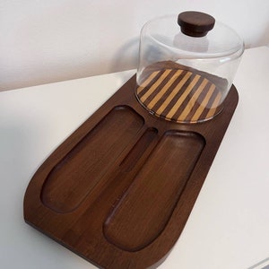 Puede incluir: Una bandeja de servir de madera con una cúpula de vidrio y un inserto de madera a rayas. La bandeja tiene dos compartimentos y una pequeña ranura en el centro. La cúpula tiene un pomo de madera en la parte superior.