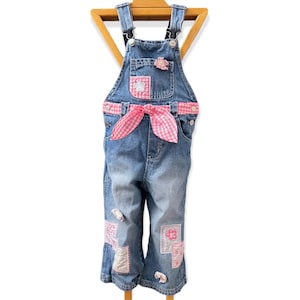 May include: Blue denim dungarees with pink gingham details and patches. The dungarees have a pink gingham bow at the waist and decorative patches with floral and geometric designs. A small pink flower decorates the bib pocket.