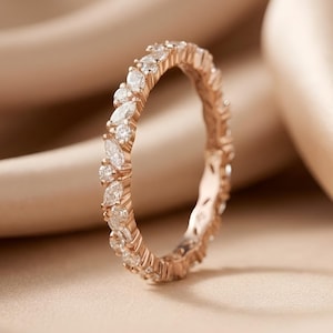 May include: A rose gold eternity ring with alternating marquise and round-cut clear gemstones. The ring is set against a soft, neutral-toned background, highlighting the sparkle of the stones. A delicate piece of jewelry.