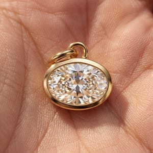 May include: An oval-shaped pendant featuring a faceted, clear, colourless gemstone set in a polished yellow gold bezel. The pendant includes a small, circular gold jump ring at the top for attachment to a chain, showcasing a brilliant cut design.