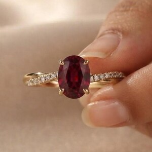 Peut inclure: Une bague en or avec une pierre précieuse rouge foncé de taille ovale fixée par quatre griffes. L'anneau présente un design torsadé orné de petites pierres claires serties pavé le long de la tige, offrant une allure raffinée et élégante.