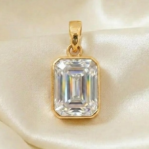 Puede incluir: Un colgante de oro con una gema grande, rectangular y transparente. La gema está engastada en un bisel de oro y cuelga de una fianza de oro. El colgante se muestra sobre una tela sedosa de color crema.