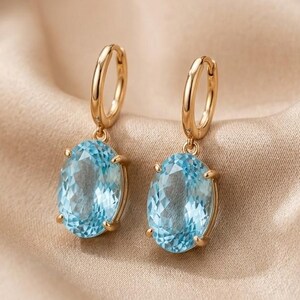 May include: A pair of gold hoop earrings featuring oval-cut, light blue faceted gemstones suspended from each hoop. The earrings are crafted with a polished gold metal finish, showcasing a classic drop design with clear, bright blue stones.