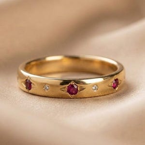 Puede incluir: Un anillo de oro con tres piedras preciosas de color rubí y dos piedras claras más pequeñas. El anillo está sobre una tela beige suave. Las piedras están engastadas en forma de estrella.