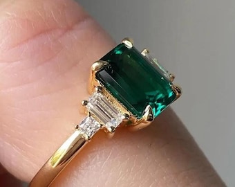 Op de afbeelding: Een gouden ring met een grote smaragdgeslepen groene edelsteen en twee kleinere rechthoekige witte edelstenen. De ring heeft een gepolijste gouden band.