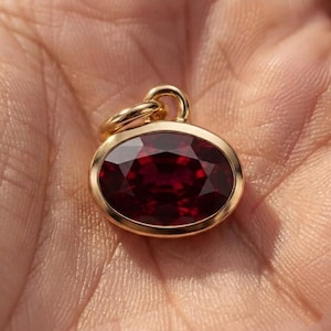 Könnte beinhalten: Ein Goldanhänger mit einem oval geschliffenen, tiefroten Edelstein in einer polierten Goldfassung. Der Anhänger verfügt über eine kleine Goldöse an der Oberseite zur Befestigung und zeigt eine facettierte Oberfläche, die das Licht reflektiert.