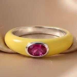 Könnte beinhalten: Ein Ring mit einer glatten, hellgelben Emaille-Schiene und einer polierten, silberfarbenen Innenseite. In der Mitte der Schiene befindet sich ein einzelner, ovaler, leuchtend fuchsia-pinkfarbener Edelstein in einer einfachen silberfarbenen Fassung.
