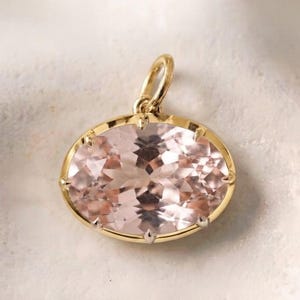May include: An oval-shaped, pale pink gemstone pendant set in a polished yellow gold frame. The gemstone features a faceted cut that reflects light, and the pendant includes a small, circular gold bail at the top for attachment to a chain.