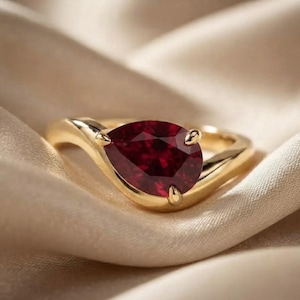 May include: A gold ring featuring a single pear-shaped deep red gemstone set in a prong setting. The band has a smooth, polished finish and a curved design that tapers toward the central stone, creating a continuous and elegant look for the piece.