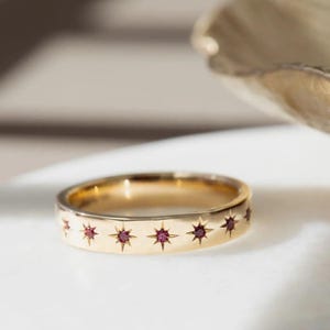 Pode incluir: Um anel de ouro com uma banda com um padrão repetitivo de estrelas, cada uma contendo uma pequena pedra preciosa vermelha e redonda. O anel é colocado sobre um fundo macio e neutro, destacando seu acabamento polido e design intrincado.
