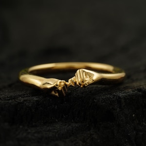 Könnte beinhalten: Ein goldener Ring mit zwei ineinander verschlungenen Händen, die einen Kreis bilden. Der Ring liegt auf einem dunklen Hintergrund.