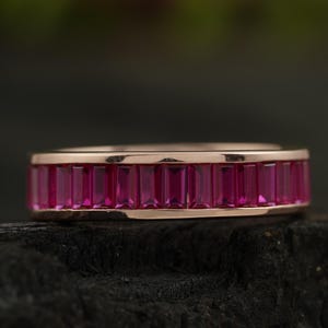 Puede incluir: Un anillo de banda de oro rosa con una fila de piedras preciosas rosadas en forma de baguette.