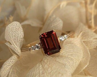Könnte beinhalten: Ein goldener Ring mit einem großen, rechteckigen, tiefroten Edelstein. Der Ring ist mit kleineren, klaren Edelsteinen auf beiden Seiten des Hauptsteins verziert, vor einem weichen, neutralen Hintergrund.