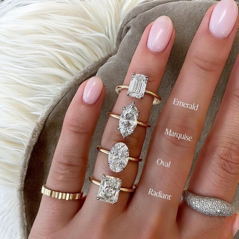 K&ouml;nnte beinhalten: Eine Auswahl an Goldringen mit verschiedenen Diamantschliffen: Smaragd, Marquise, Oval und Radiant. Die Ringe werden auf einer Hand mit rosa Nagellack pr&auml;sentiert. Text kennzeichnet den Schliff jedes Rings. Ein strukturierter Goldring und ein diamantbesetzter Ring sind ebenfalls zu sehen.