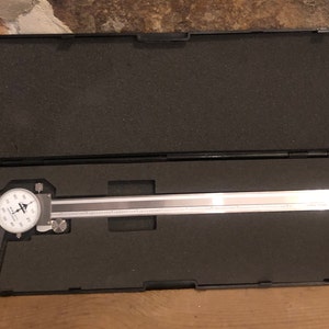 May include: A silver metal caliper with a black case. The caliper has a dial with markings in inches and a sliding scale. The case is made of black plastic and has a foam lining.