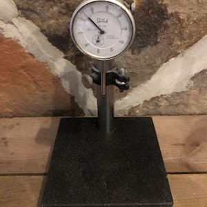 May include: A black dial dial indicator with a white face and a black base. The dial indicator is mounted on a black granite base. The dial indicator reads "Telsch" and has a range of 0 to 100.