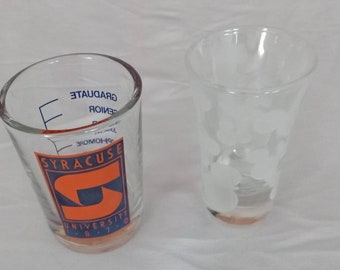 May include: Two clear glass shot glasses. The one on the left has a blue and orange Syracuse University logo and markings for measuring liquid. The one on the right has a frosted design.