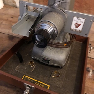 May include: A vintage TDC Selectron 35mm slide projector in a brown leather case. The projector is silver and gray with a black lens. The case has a brass latch and a warning label that reads "Do not operate projector in case."