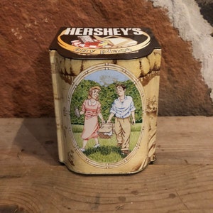 May include: A vintage tin box with a brown lid and a yellow base. The lid features the Hershey's logo and the text "Simply Irresistible". The base features a cartoon illustration of a couple on a picnic in a green field.
