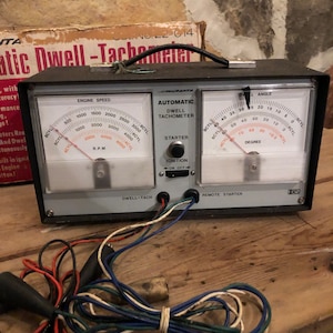 May include: A black and silver automotive dwell tachometer with three gauges. The gauges measure engine speed in RPM, dwell angle in degrees, and ignition timing. The device has a starter button and an on/off switch. The device is connected to a set of wires.