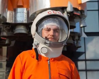 Pode incluir: Uma pessoa vestindo um capacete de astronauta branco com o texto "CCCP" no lado e um macacão laranja, em pé na frente de um foguete.