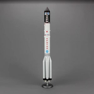 May include: A white and black model of a Proton rocket with the word "PROTON" in red Cyrillic letters and the word "ЗАРЯ" in black Cyrillic letters on the side. The rocket is on a black stand.