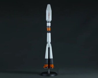Pode incluir: Um modelo de foguete branco e laranja com a palavra "Soyuz" escrita no lado. O foguete está em um suporte preto com três pernas.