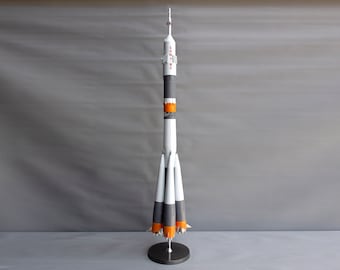 Pode incluir: Um modelo de foguete branco e laranja com o texto "СОЮЗ" no lado. O foguete está em um suporte preto.