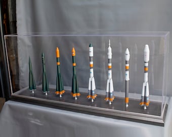 Pode incluir: Uma vitrine com oito maquetes de foguetes de diferentes tamanhos e cores. Os foguetes estão dispostos em uma fila do menor para o maior. Todos os foguetes são brancos com detalhes em laranja e verde.