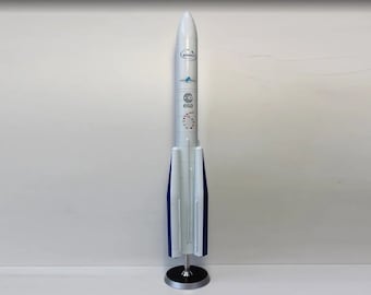 Pode incluir: Um modelo de foguete branco com detalhes azuis, sobre uma base prateada. O foguete apresenta logotipos e texto impressos, incluindo "Ariane" e "esa". O design é aerodinâmico, indicativo de viagens espaciais, com cerca de 60 cm de altura.