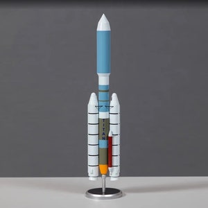 Może przedstawiać: Model rakiety Titan w kolorze niebieskim, białym i zielonym. Rakieta stoi na srebrnym stojaku. Na rakiecie widnieje napis "TITAN".