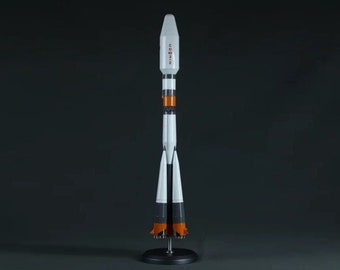 Pode incluir: Um modelo de foguete branco e laranja com a palavra "Soyuz" escrita no lado. O foguete está em um suporte preto com três pernas.
