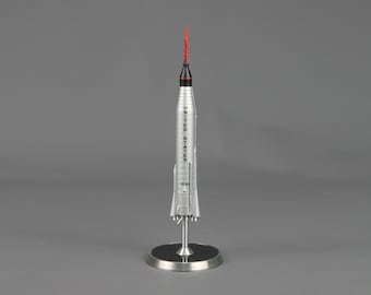 Pode incluir: Um modelo de foguete prateado com ponta vermelha e as palavras "UNITED STATES" impressas verticalmente. O foguete é montado em um suporte prateado com base preta. Modelo de foguete Mercury-Redstone.