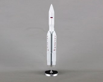 Pode incluir: Um modelo branco do foguete Angara A5 com a bandeira russa no topo. O foguete está em um suporte prateado com o texto "АНГАРА-А5" no lado.