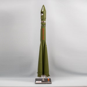 May include: A green model rocket with the word "СССР" in red letters on the side. The rocket is on a black stand with a small figure of a person standing next to it.